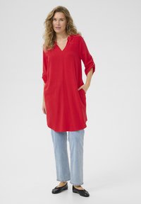 Röd tunika med V-ringning och trekvartsärmar, utformad med fickor; kombinerad med ljusblå raka jeans och svarta ballerinas.