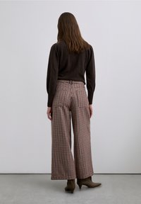 Pulover maro, pantaloni largi în carouri roșii, albe și maro și cizme maro cu toc la gleznă. Textura pare moale; designul include buzunare mari.