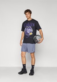 Camiseta negra con gráfico púrpura de "Lakers", combinada con pantalones cortos de malla grises. El modelo sostiene un baloncesto, vistiendo zapatillas y calcetines negros.