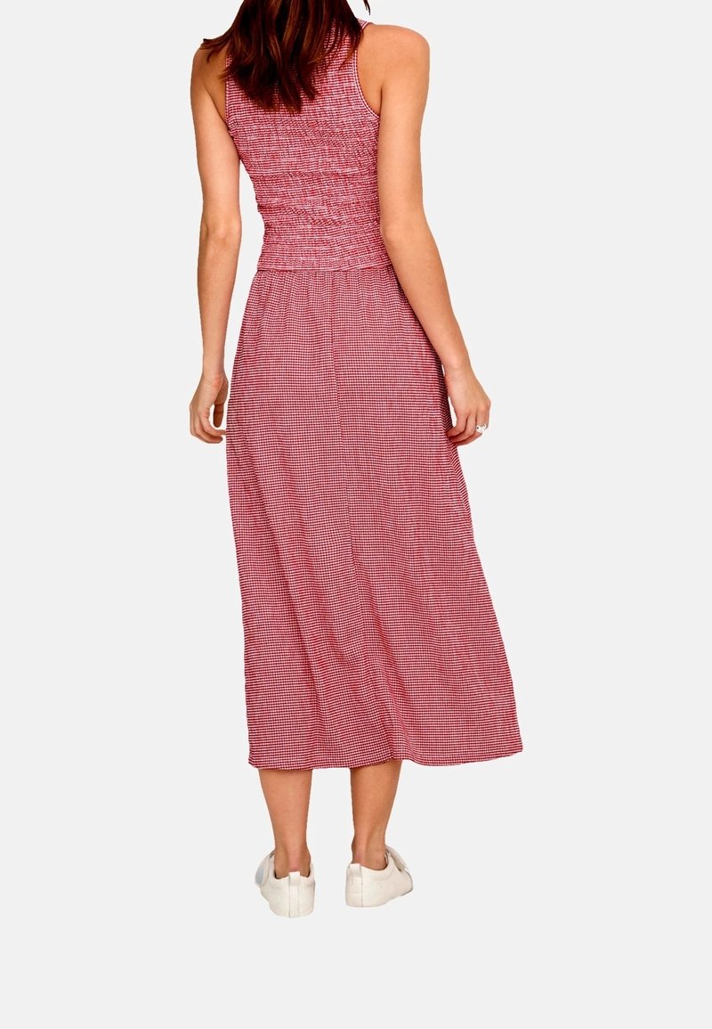 Femme debout, de dos, portant une robe rouge et blanche sans manches, longue jusqu'aux chevilles, et des baskets blanches sur un fond uni.