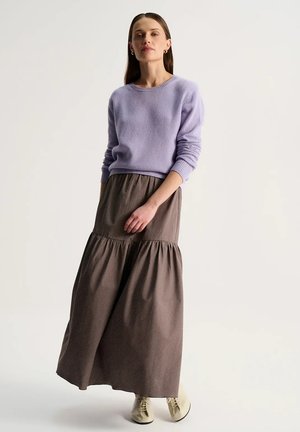 Kobieta ubrana w lawendowy sweter, brązową marszczoną spódnicę maxi i kremowe kozaki wiązane, stoi opierając jedną rękę na spódnicy.