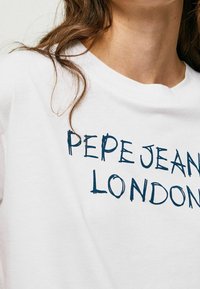 Camiseta blanca de algodón con cuello redondo, que presenta el texto gráfico azul "PEPE JEANS LONDON" en el frente. Textura suave visible.