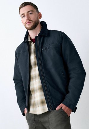 Navy blue jacket with a zip front, high collar, and a chest pocket. Worn over a beige plaid shirt, complemented by gray pants.