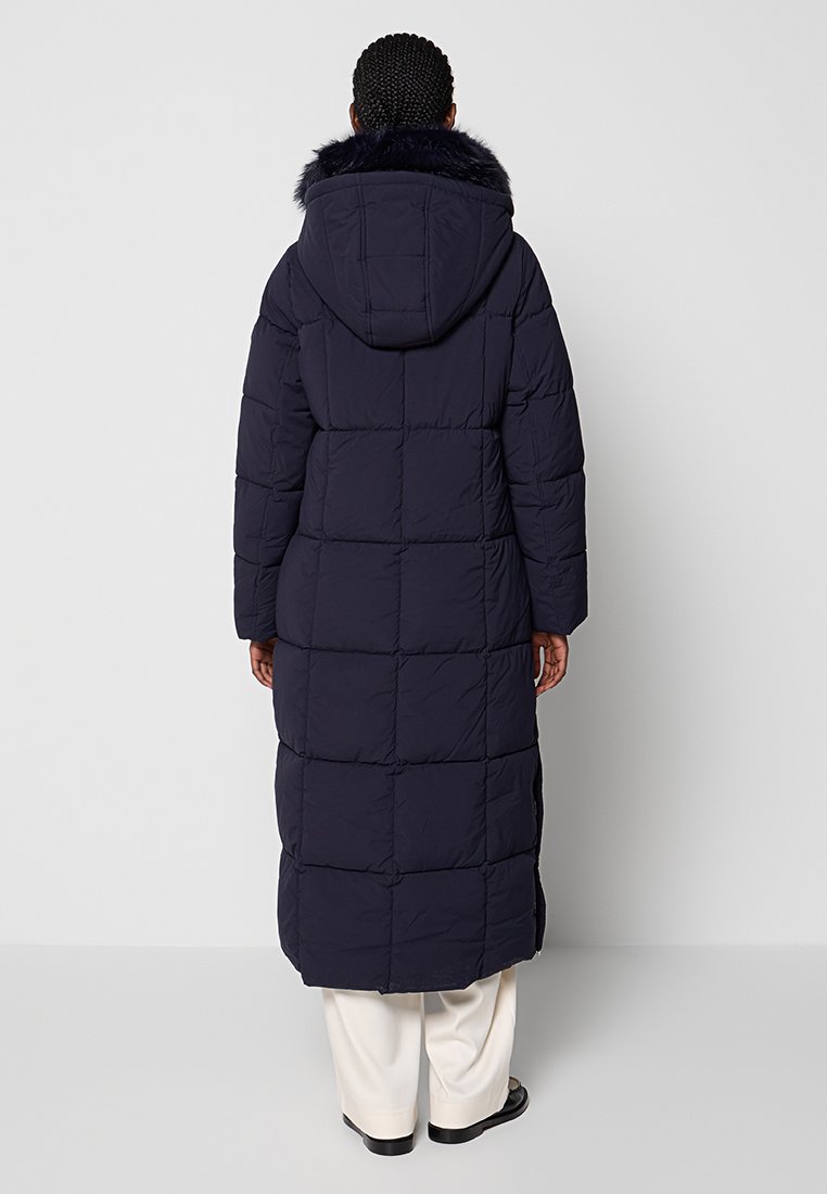 Lungo cappotto imbottito blu navy con cappuccio; presenta un colletto in pelliccia sintetica, una texture trapuntata e spacchi laterali per facilità di movimento.