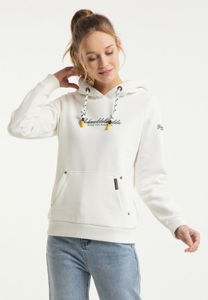 Jonge vrouw met licht haar in een paardenstaart, draagt een witte hoodie met tekst en een blauwe spijkerbroek, poseert met één hand achter haar hoofd.