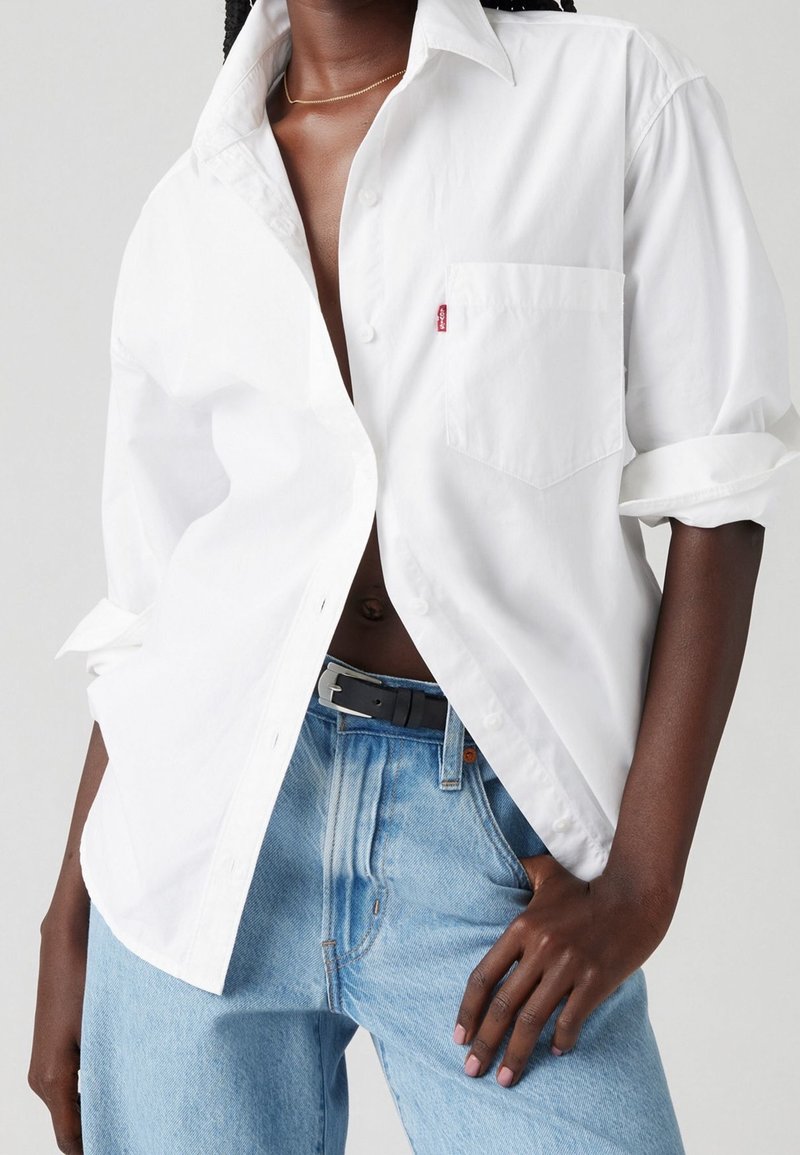 Personne portant une chemise blanche à col déboutonnée avec une poche poitrine visible, un jean bleu clair et une ceinture noire, la main posée sur la hanche.