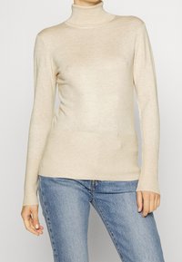 Beige Rollkragenpullover aus weichem, geripptem Strick, figurbetontes Design mit langen Ärmeln und glatter Oberfläche, kombiniert mit hellblauen Jeans.