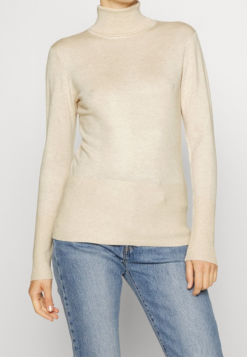 Beige Rollkragenpullover aus weichem, geripptem Strick, figurbetontes Design mit langen Ärmeln und glatter Oberfläche, kombiniert mit hellblauen Jeans.