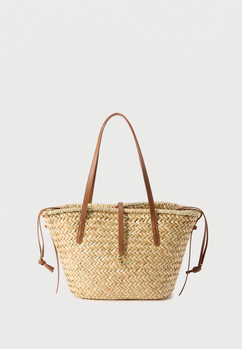 Πλεκτή τσάντα ώμου από ψάθα με μακριές καφέ δερμάτινες λαβές, δερμάτινο λουράκι με χρυσό κούμπωμα και πλαϊνά κορδόνια στήριξης σε ανοιχτό φόντο.