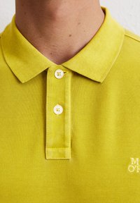 Limonkowa koszulka polo z fakturowanej tkaniny, z dwoma guzikami i kołnierzykiem. Posiada białe, haftowane logo na dolnym lewym rogu.