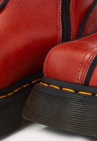 Gros plan sur des bottes en cuir rouge avec des semelles côtelées noires, des surpiqûres jaunes et une fermeture éclair noire sur le côté.