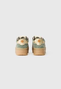 Une paire de baskets en suede vert avec des accents beiges et des semelles en caoutchouc, vue de l'arrière sur un fond blanc.