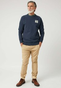 Hombre con cabello y barba canosos, vistiendo un sudadera azul marino con el texto "Sueña en Grande", pantalones beiges y botas marrones, de pie con las manos en los bolsillos.