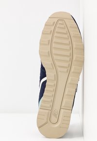 Semelle d'une basket bleu marine à semelle beige avec grip texturé et logo "NB" sur le talon, inclinée verticalement contre un fond blanc d'angle.