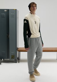 Cream and green long-sleeve athletic top with zip, gray sweatpants, beige shoes, curly hair, posed by lockers and bench.