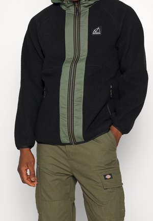 Man draagt een zwart fleecejack met een groen ritsdetail en olijfgroene cargobroek met zakken aan de zijkant.