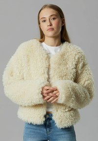 Nepbont jas in off-white, met een boxy silhouet, brede mouwen en een zachte, gestructureerde textuur. Gelaagd over een witte top en blauwe jeans.