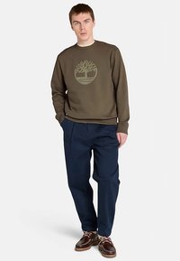 Felpa verde oliva con logo circolare di un albero, abbinata a pantaloni svasati blu scuro e scarpe da barca marroni con dettagli in pizzo.