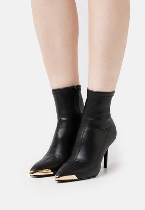 Bottes en cuir noir montant à la cheville, avec un bout pointu, des accents dorés à l'extrémité et une silhouette élégante dotée d'une fermeture éclair latérale.