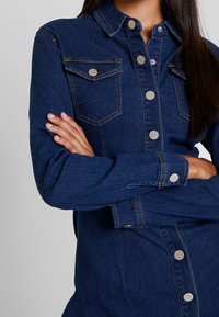 Mörkblå denimskjorta med krage, silverfärgade tryckknappar och framfickor. Tyget har en slät, strukturerad yta.