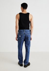 Young man standing facing away wearing black tank top, blue denim jeans with back pockets, and black shoes on plain light background.