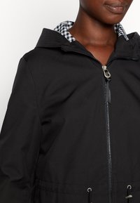 Femme portant une veste noire zippée avec doublure de col à carreaux, visible des épaules au menton sur fond blanc.