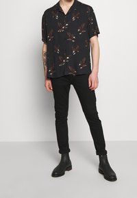 Mann in schwarzem kurzärmeligen Hemd mit Adlerdruck, schwarzen Slim-Fit-Hosen und schwarzen Knöchelstiefeln, der vor einem schlichten Hintergrund steht.