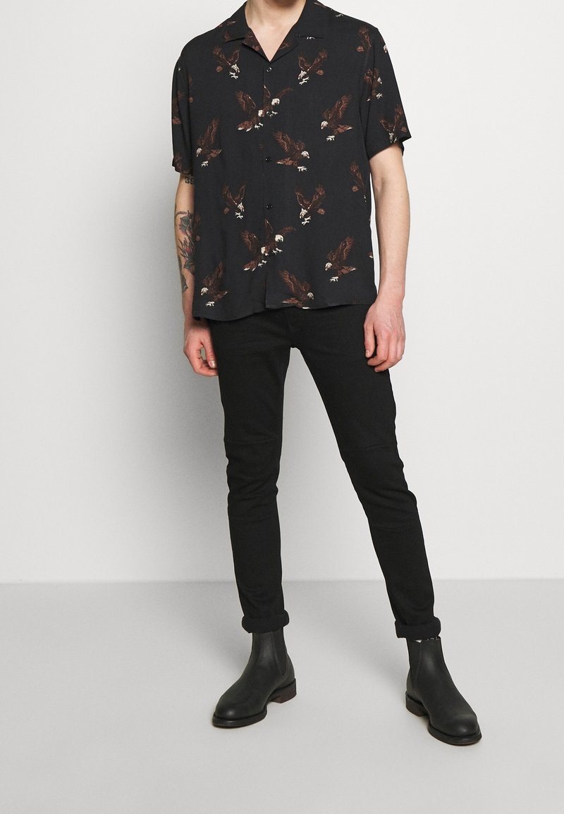 Mann in schwarzem kurzärmeligen Hemd mit Adlerdruck, schwarzen Slim-Fit-Hosen und schwarzen Knöchelstiefeln, der vor einem schlichten Hintergrund steht.