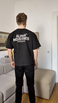 Camisa preta de manga curta com um padrão texturizado, apresentando o texto branco "ALPHA INDUSTRIES STUDIO EDITION" nas costas.