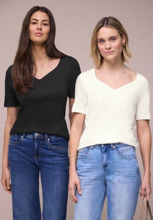 Zwei Frauen stehen nebeneinander, eine in schwarzem kurzärmligem Oberteil und dunklen Jeans, die andere in weißem kurzärmligem Oberteil und hellblauen Jeans.