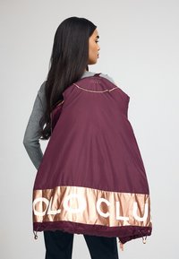 Marin puffer-väst med bronsaccenter, med en glänsande remsa med texten "POLO CLUB", dragkedja fram och dragsko i nederkanten för justering.