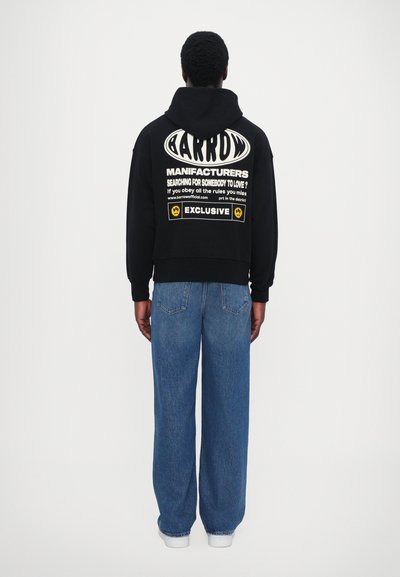 Haut à capuche noir avec un texte graphique blanc dans le dos, associé à un jean en denim bleu ample et des baskets blanches. Le texte comprend des illustrations et des slogans.