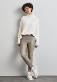 Vit fluffy tröja med hög krage, tillsammans med ljusbruna åtsittande byxor. Vita sneakers med beige detaljer. Lockigt hår.