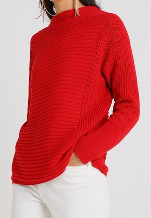 Femme portant un pull en maille côtelée rouge vif à manches longues et un pantalon blanc, main dans la poche, sur un fond clair uni.