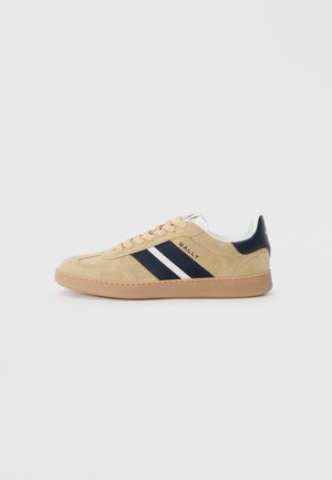 Beige suede sneakers with navy and white stripes, round toe, and a tan rubber sole. Features lace-up closure and branded detail on the side.