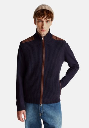 Chaleco con zipper en un oscuro color navy con un frente de punto texturizado y detalles en marrón en los hombros. Mangas largas en tejido suave. La modelo lleva un gorro de punto beige.