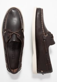 Braune Lederbootsschuhe mit weißen Gummisohlen, verziert mit dekorativen Nähten und Schnürung. Die Innenauskleidung ist dunkel mit Branding-Details.