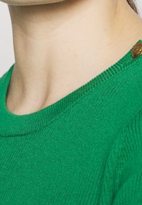 Vihreä neulottu neulepaita pyöreällä pääntiellä, jossa on teksturoidu pinta ja tunnusomainen ruskea nappidetalji olkapään lähellä.