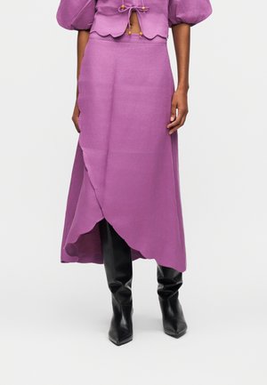 Femme portant une jupe portefeuille violette et un haut assorti à manches bouffantes, associée à des bottes noires à bouts pointus montant jusqu'au genou, debout devant un fond blanc.