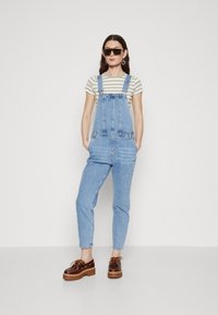 Pantalones de mezclilla azul claro con un corte suelto, que cuentan con tirantes ajustables y bolsillos frontales, combinados con una camisa a rayas y zapatos de plataforma marrones.