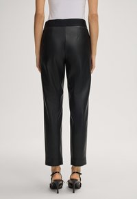 Woman wearing black high-waisted leather pants with a black branded waistband and black high-heeled sandals, standing facing away.