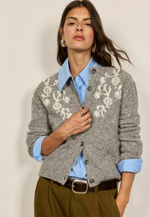 Frau trägt grauen Cardigan mit floralem Muster über einem blauen Hemd, braune Hose und goldene Ringe, posiert mit einer Hand nahe der Brust.