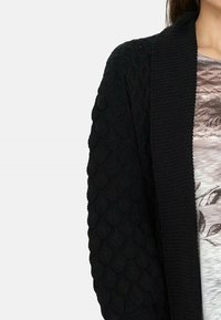 Cárdigan de punto negro con un patrón texturizado en las mangas y un cuello acanalado, que ofrece un aspecto acogedor y cálido.