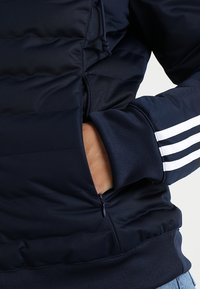Námořnická puffer bunda se vodorovnými �švy, zipovými kapsami a bílými třemi pruhy na rukávech. Hladký povrch, neformální design.