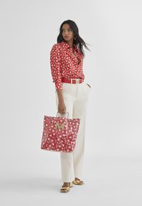 Blusa a righe rosse, pantaloni bianchi e una borsa a tracolla trasparente con un logo dorato. Sandali piatti dorati. Sfondo minimalista.