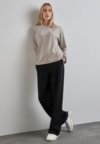 Maglione grigio chiaro a maglia con maniche lunghe, abbinato a pantaloni neri a gamba larga e sneaker bianche, su uno sfondo di parete grigia uniforme.