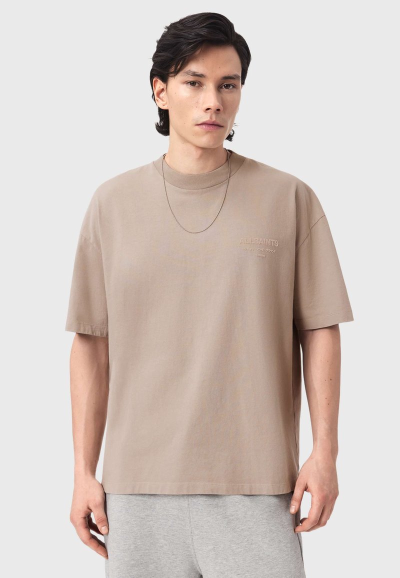 Μπεζ κοντομάνικο μπλουζάκι από απαλό βαμβάκι με άνετη γραμμή, διαθέτοντας ένα διακριτικό ανάγλυφο λογότυπο στο στήθος και λαιμόκοψη τύπου μπλούζας.