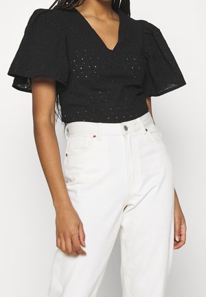 Vrouw draagt een zwarte korte mouwen cropped top met oogjes en pofmouwen, en een witte high-waisted jeans, staand tegen een witte achtergrond.