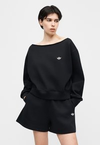 Άτομο που φοράει μαύρο φούτερ Adidas και αντίστοιχα σορτς με λευκά λογότυπα, με τα χέρια στις τσέπες, κοιτάζοντας προς τα πλάγια σε απλό φόντο.
