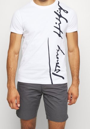 Homme portant un t-shirt blanc à manches courtes avec un texte vertical en écriture noire et un short gris foncé.
